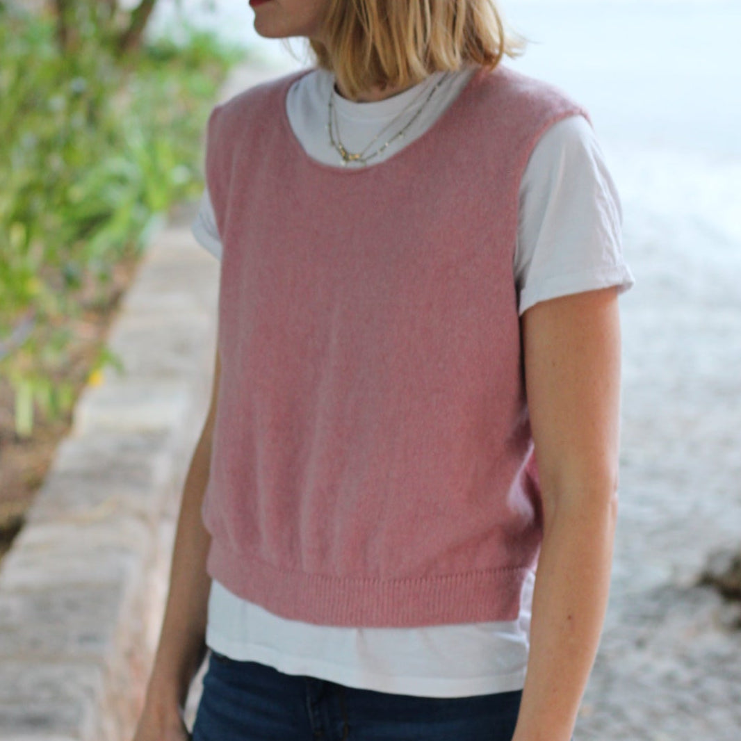 Person wearing a pink sleeveless top and blue jeans standing outdoors with greenery in the background