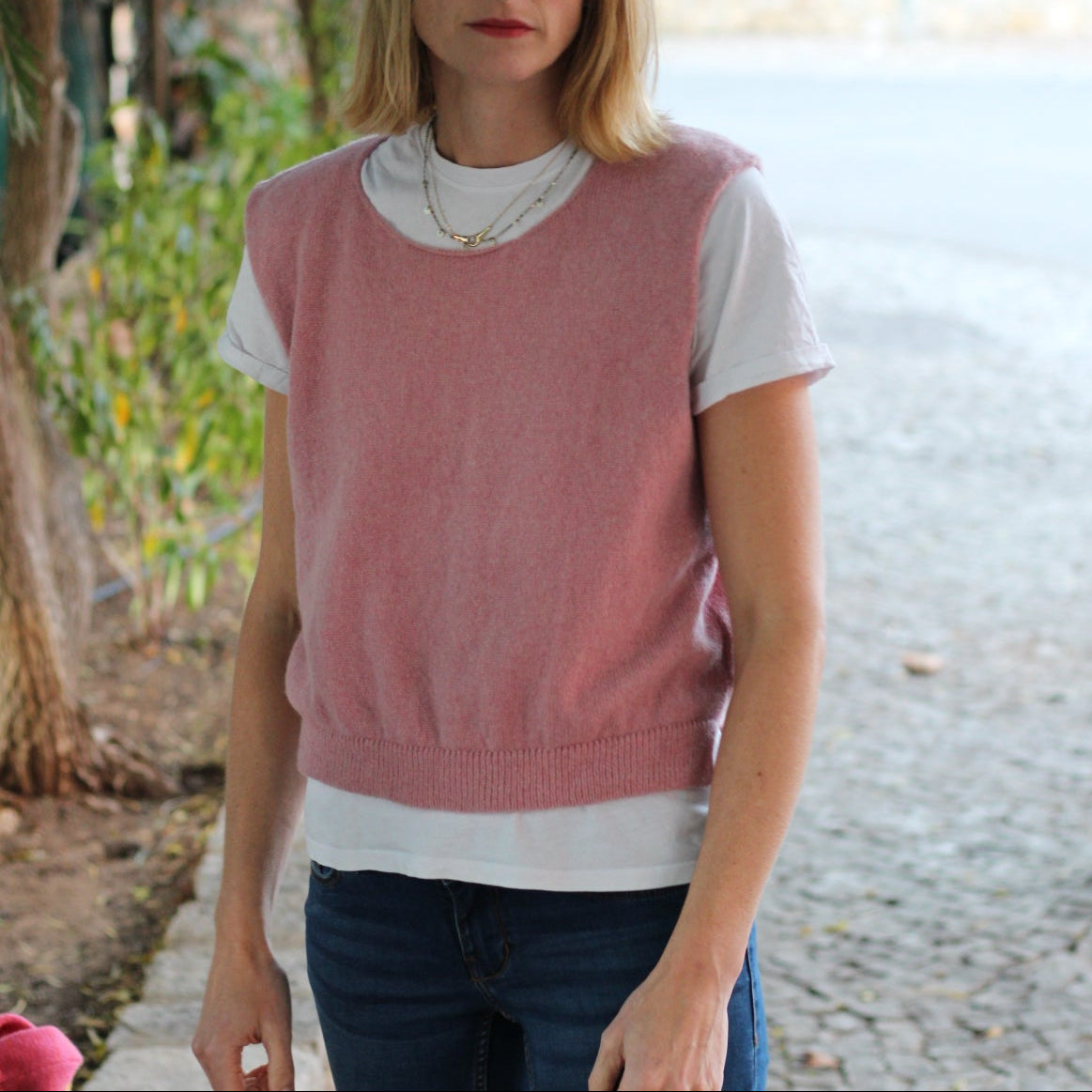 Woman wearing a pink sleeveless top and blue jeans standing on a stone path with greenery in the background