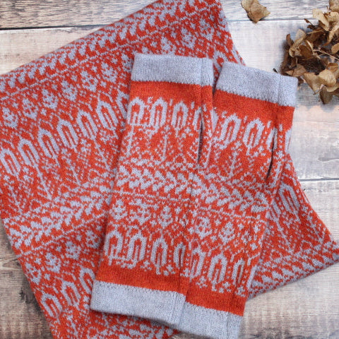Red and gray patterned scarf and gloves on a wooden surface with dried leaves.