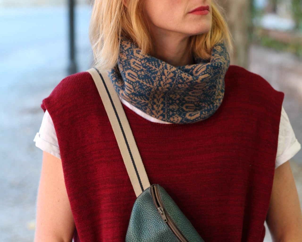Woman wearing a red vest and patterned scarf with a green bag, standing outdoors.