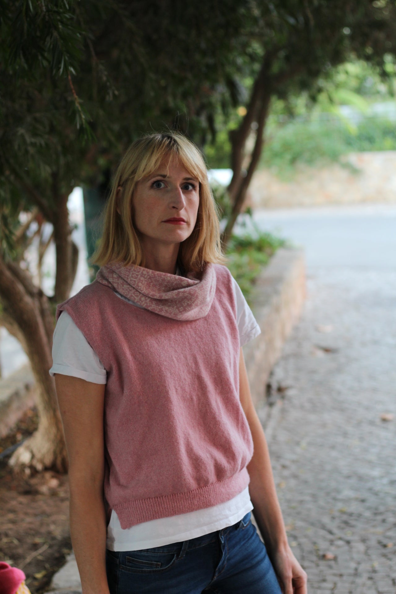 Woman wearing a pink sleeveless top and blue jeans standing outdoors with trees in the background