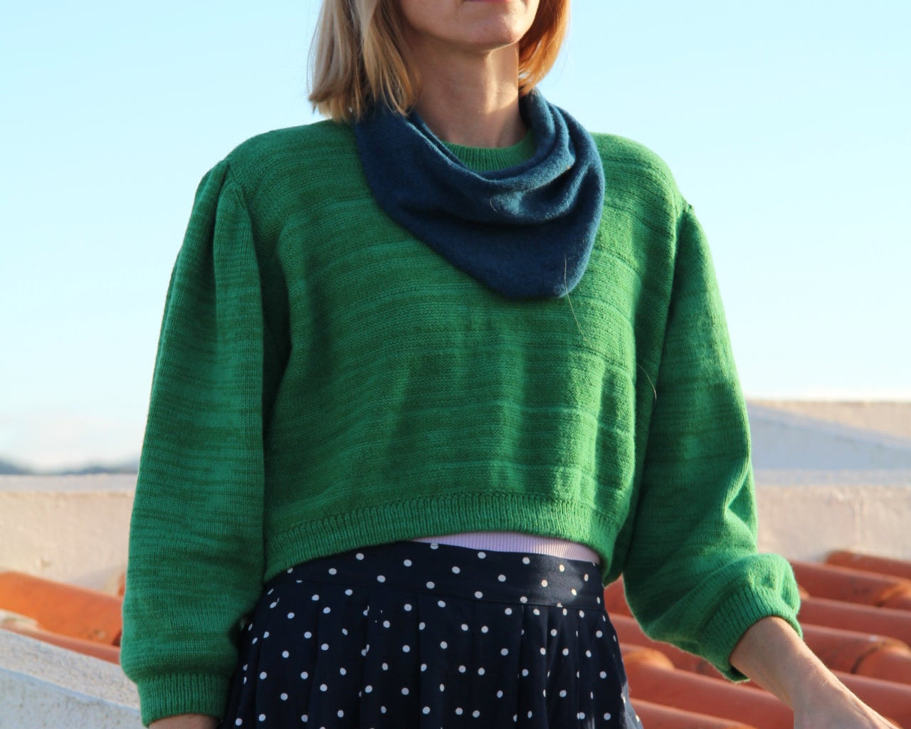 Woman wearing a green sweater and black polka dot dress against a blue sky.